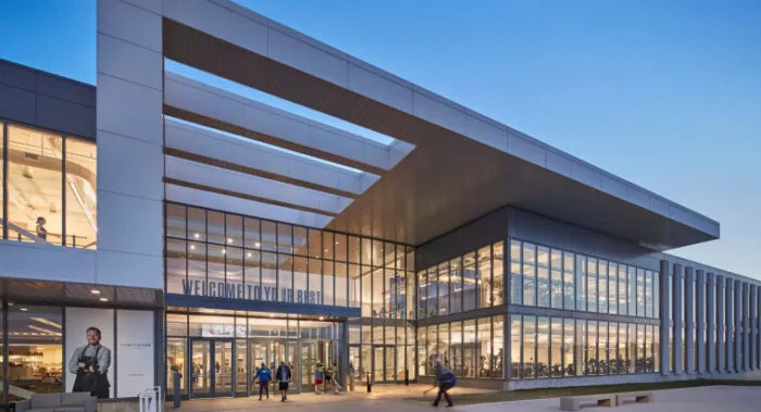 Modern glass and steel building with large windows, people walking outside, and well-lit interior visible, likely a gym or fitness center, pictured at dusk with a clear sky.