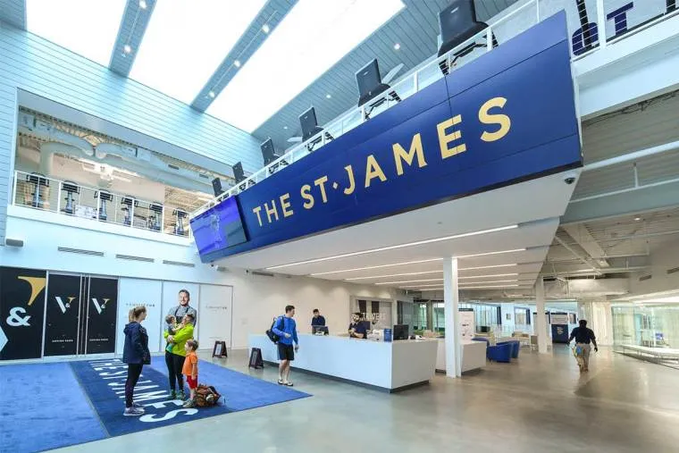 Spacious lobby of The St. James sports complex, featuring a modern blue and white design. People, including children and adults in athletic clothing, stand near the reception desk and walk through the main entrance area.