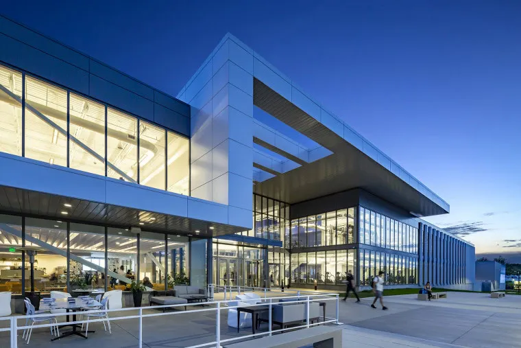 Modern building with large glass windows and geometric architecture, illuminated in the evening. People are walking near the entrance, and outdoor seating is visible on the patio in the foreground.