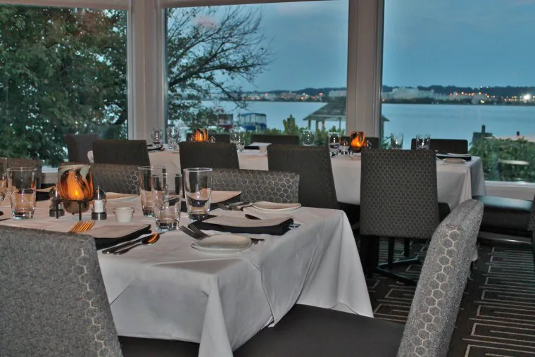 Elegant restaurant dining area with tables set with white tablecloths, plates, glasses, and napkins. Large windows offer a scenic view of water, trees, and a gazebo outside at dusk.