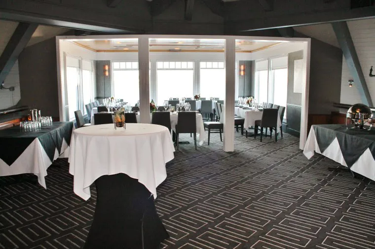 A banquet room with tables covered in white tablecloths, set for an event. Chairs surround the tables, and large windows let in natural light. The room has geometric patterned carpet and neatly arranged dining settings.