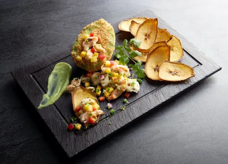 A rectangular black slate plate with plantain chips, a dollop of green sauce, ceviche topped with diced mango and vegetables, and a breaded, fried avocado half, garnished with fresh herbs.