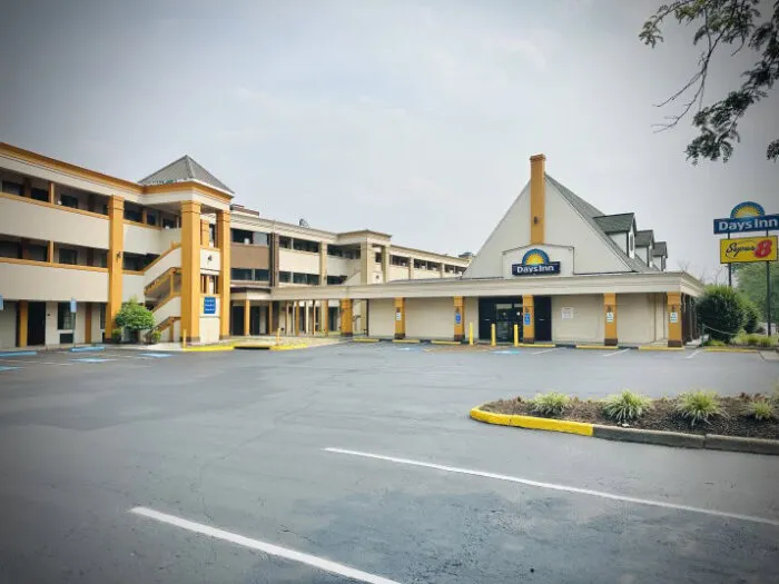 A Days Inn hotel with a large parking lot and minimal cars, featuring yellow trim and two stories, under a clear sky with some greenery around the lot and a sign visible near the entrance.