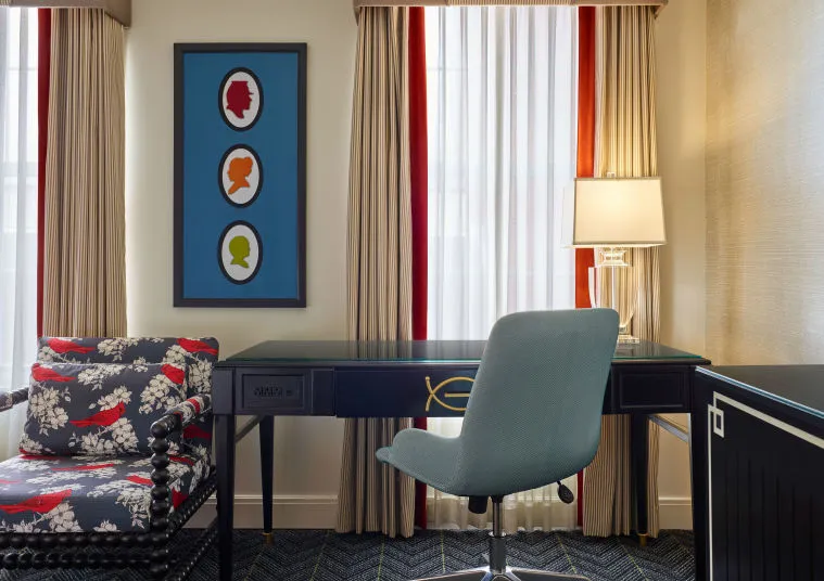 A modern hotel room features a black desk with a blue chair, a lamp, and a floral-patterned armchair. Large windows with beige curtains and a wall art with colorful silhouettes complete the decor.