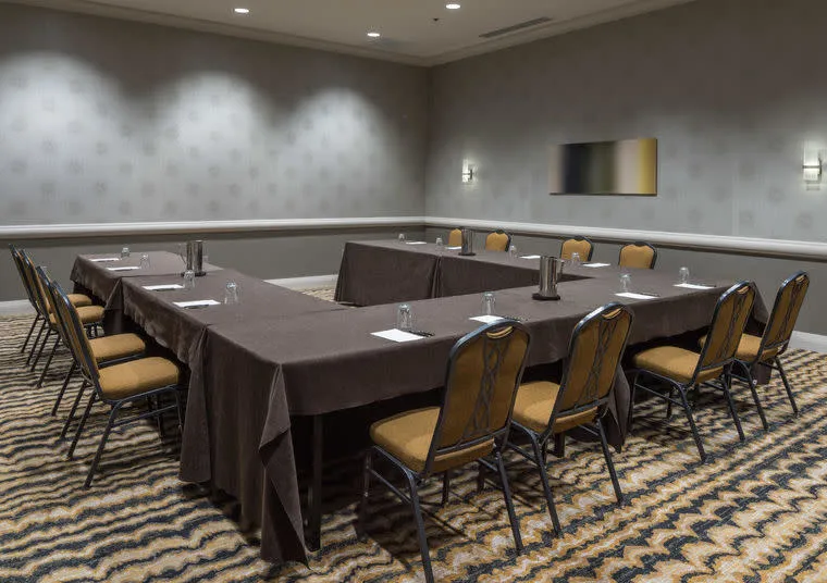 A conference room with U-shaped tables covered in brown tablecloths, surrounded by tan chairs. Each table has a water pitcher and glasses, with patterned carpet and neutral walls in the background.