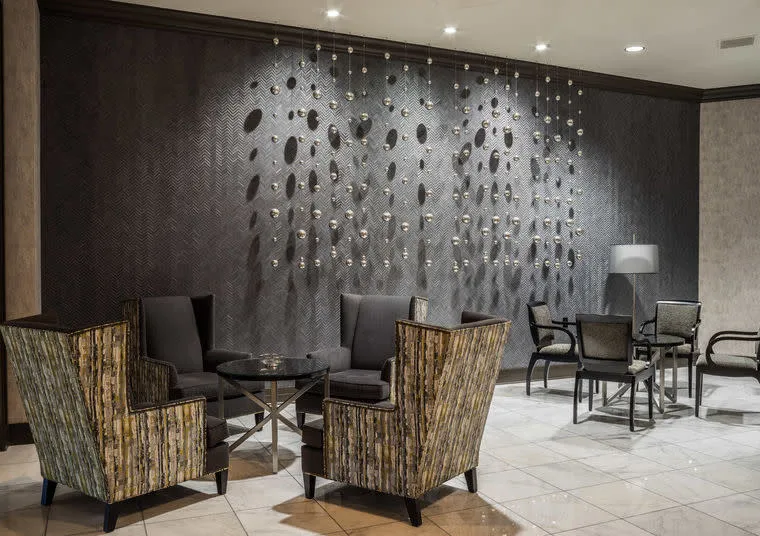 Modern hotel lobby with patterned armchairs around a glass coffee table, sleek gray chairs at a nearby dining table, tiled floor, and a dark textured wall with metallic decorative art and ceiling spotlights.