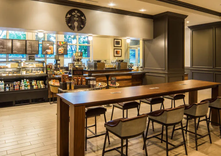 A modern Starbucks cafe interior with a long wooden table and chairs in the foreground, a counter with display cases of food and drinks, menu boards, and Starbucks logo in the background.