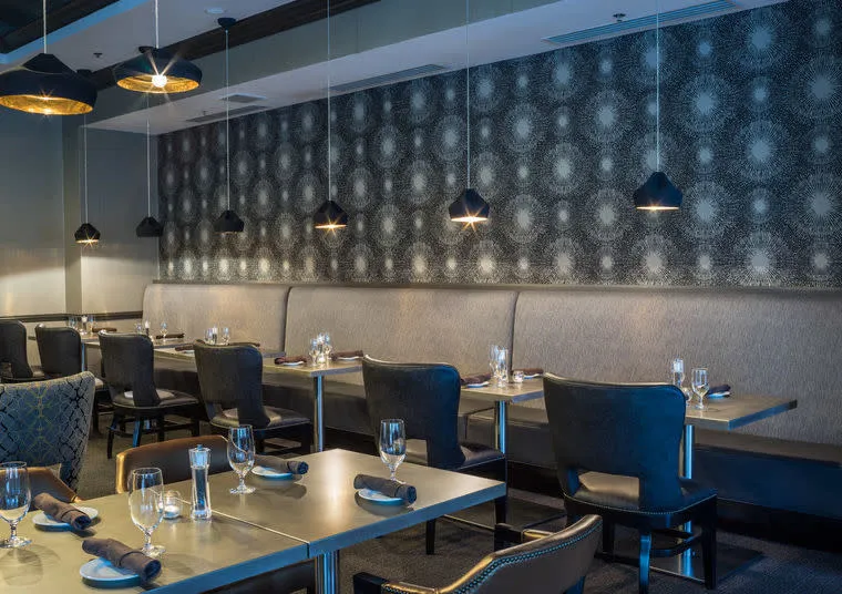 Modern restaurant interior with gray patterned wallpaper, pendant lights, cushioned bench seating, and tables set with glasses, napkins, and silverware. The ambiance is elegant and inviting.