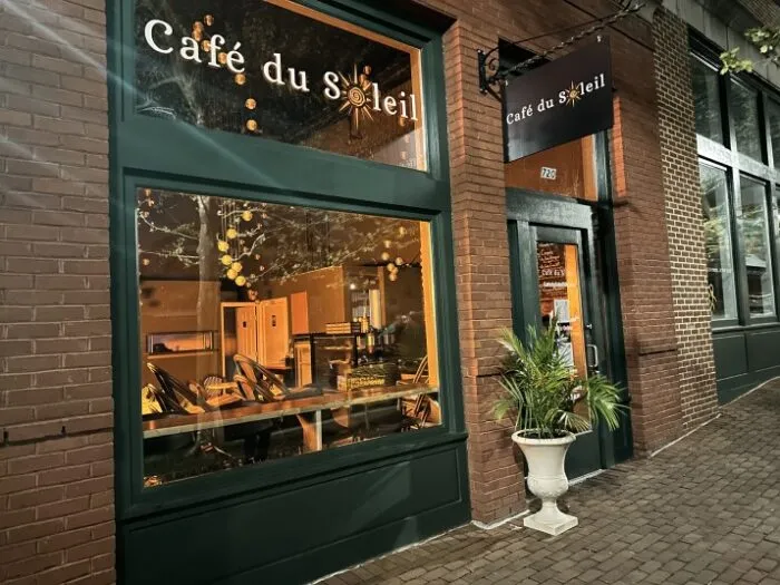 Exterior of Café du Soleil at night, with warm light glowing from inside. The green storefront features large windows, plants outside, and a hanging sign displaying the café’s name. Brick sidewalk runs along the front.