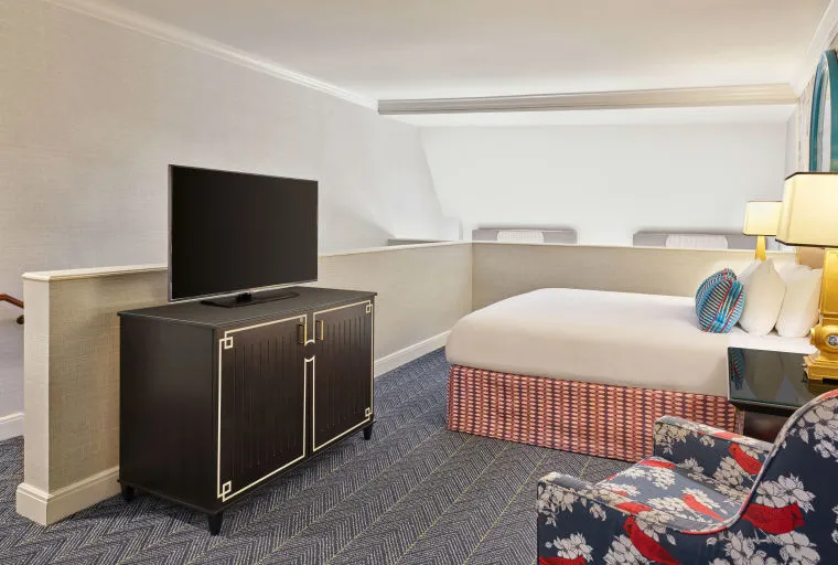 A hotel room features a bed with white linens and a patterned base, a black TV stand with a flat-screen TV, a floral armchair, and a small table with two lamps next to the bed. The room has a modern, clean design.