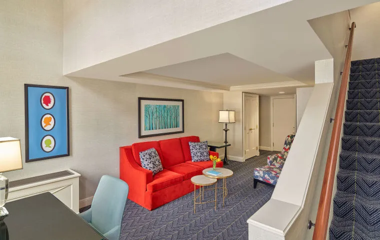 A modern hotel suite with a red sofa, patterned armchair, two round coffee tables, wall art, a desk with a blue chair, and a staircase leading upstairs. The room is brightly lit and decorated in neutral tones.