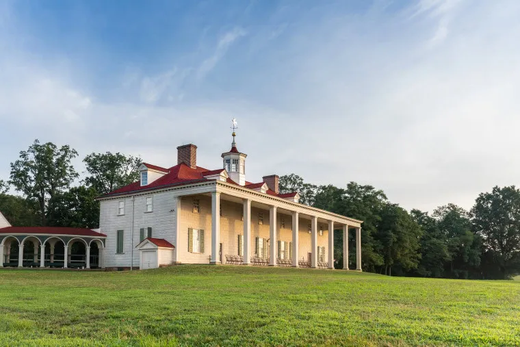 A historic white mansion with a red roof and tall columns stands on a grassy lawn, surrounded by trees under a partly cloudy sky.