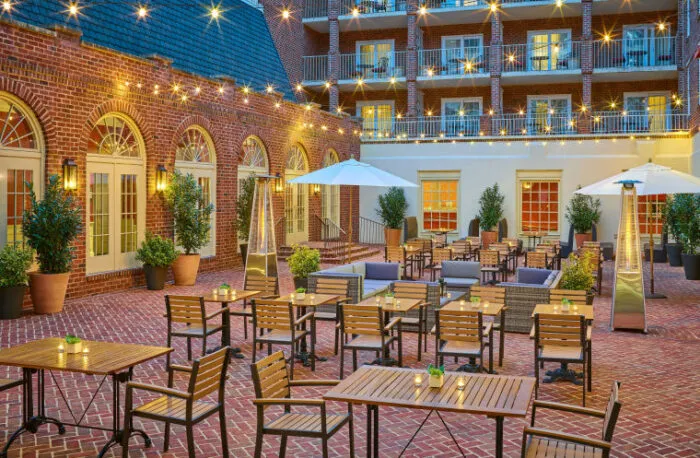 Outdoor brick courtyard with wooden tables and chairs, string lights overhead, potted plants, and white umbrellas. Cozy seating areas and a multi-story building with arched windows create a welcoming atmosphere.