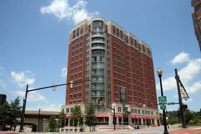 A tall red-brick apartment building with large windows and balconies stands on a street corner under a blue sky. Traffic lights, street signs, and trees are visible around the building.