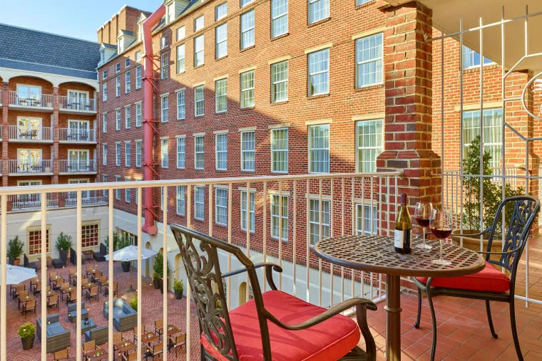 A balcony with a table, two chairs with red cushions, and a bottle and glasses of wine overlooks a courtyard with tables, umbrellas, and a brick building with many windows.