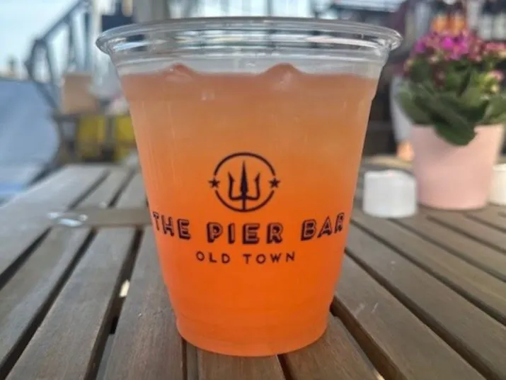A plastic cup with an orange-pink drink sits on a wooden table. The cup is labeled The Pier Bar Old Town. In the background, there is a blurred potted plant and outdoor seating.