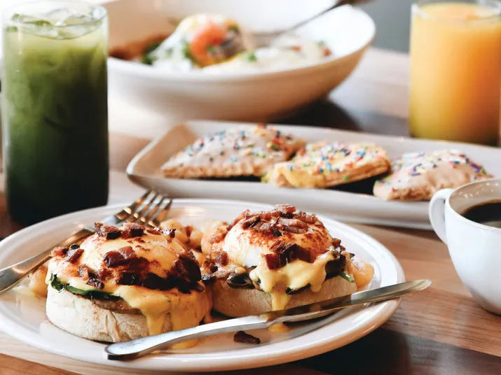 A plate of eggs Benedict with bacon, a glass of iced green drink, orange juice, coffee, homemade pastries with colorful sprinkles, and a salad bowl, all set on a wooden table for breakfast.