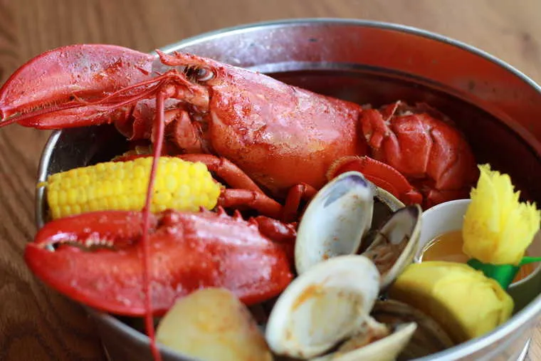 A metal bowl filled with a whole cooked lobster, clams, corn on the cob, potatoes, a lemon wedge, and a cup of melted butter on a wooden table.