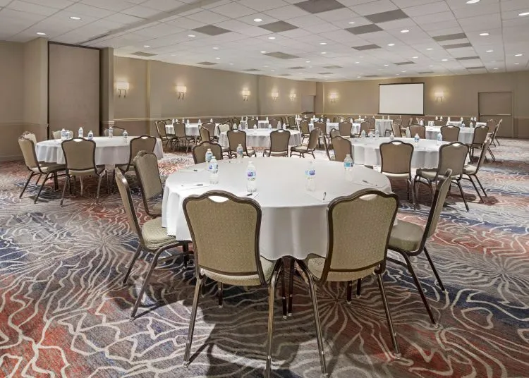 A large conference room with round tables covered in white tablecloths, each surrounded by chairs and set with bottled water and glasses. The room has patterned carpet, beige walls, and a projector screen at the front.