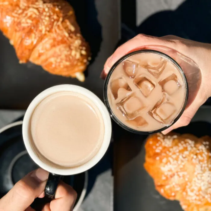 Two hands hold drinks above a table: one holds a cup of hot coffee, the other holds a glass of iced coffee. Two croissants with sesame seeds are on black plates in the background. Bright natural light shines on the scene.