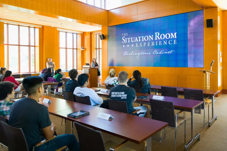 A group of students sits at desks in a bright, modern classroom, listening to a speaker near a large screen displaying The Situation Room Experience: Washingtons Cabinet.