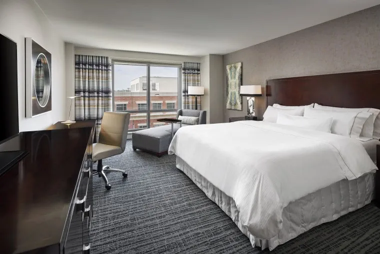 Modern hotel room with a large bed, white bedding, two bedside lamps, a desk with a chair, an armchair by the window, and a large window with a city view. Decor features neutral tones and abstract art.