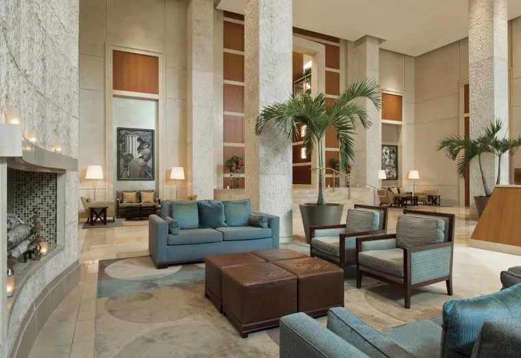 Spacious modern hotel lobby with tall ceilings, neutral walls, large pillars, blue seating, leather ottomans, potted palm trees, wall art, lamps, and a fireplace on the left.