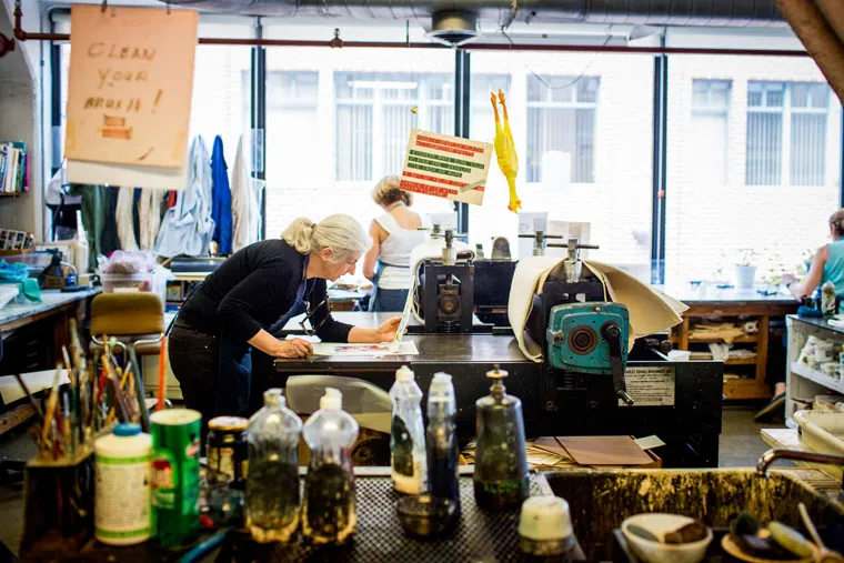 A woman with gray hair works at a table in a cluttered art studio with tools, bottles, and supplies. Two other people are visible in the background near large windows. Signs and artwork hang from the ceiling and walls.