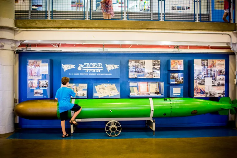 A boy in a blue shirt stands with one foot on a large green torpedo displayed in a museum. Behind him is an exhibit with photos, text, and diagrams about torpedoes, set against a blue wall.