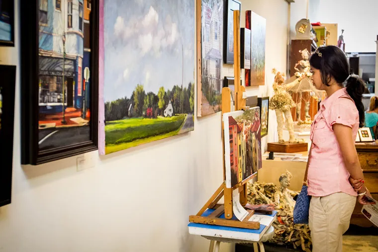 A woman in a pink shirt stands in an art gallery, closely observing various colorful paintings and sculptures displayed on the walls and easels around her.