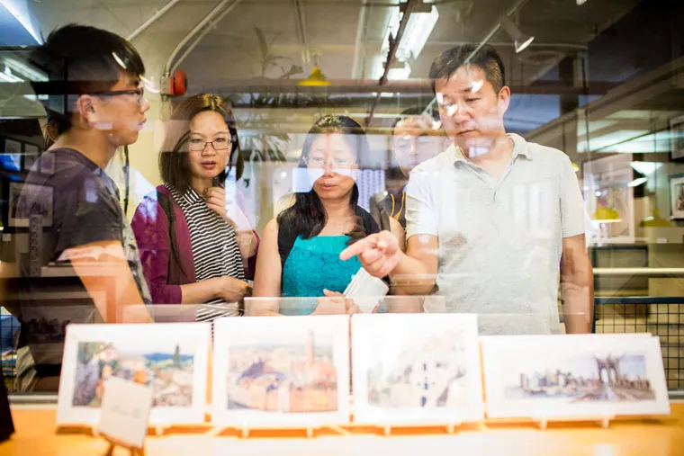 Four people stand together indoors, looking at a display of postcards or photographs behind glass; one man points at an item while reflections show lights and parts of the room.