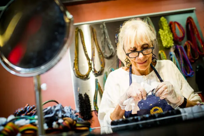 An older woman with white hair and glasses, wearing gloves and an apron, works on crafting jewelry at a counter surrounded by colorful necklaces displayed on the wall behind her.