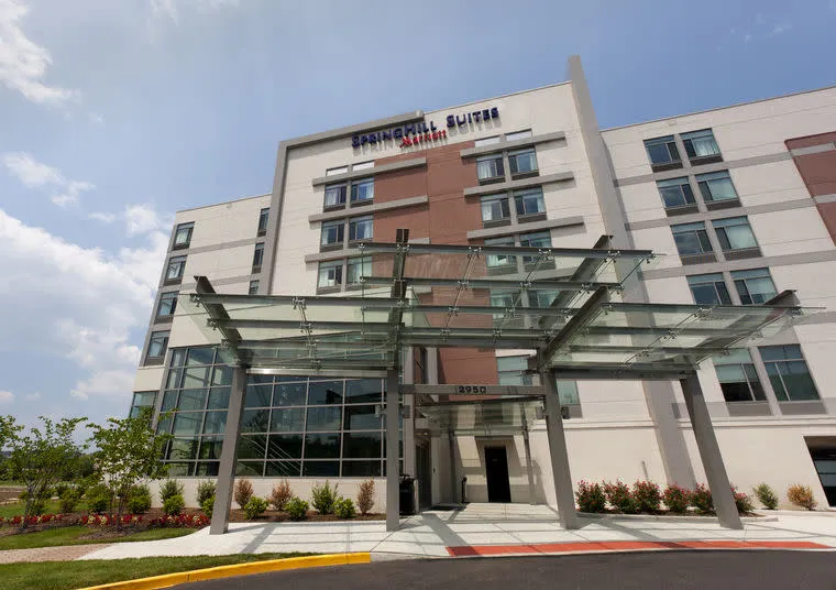Modern hotel exterior with large glass awning at entrance, beige facade, and multiple windows. The sign above reads “SpringHill Suites.” Landscaping with bushes and small trees surrounds the building. Blue sky with some clouds.