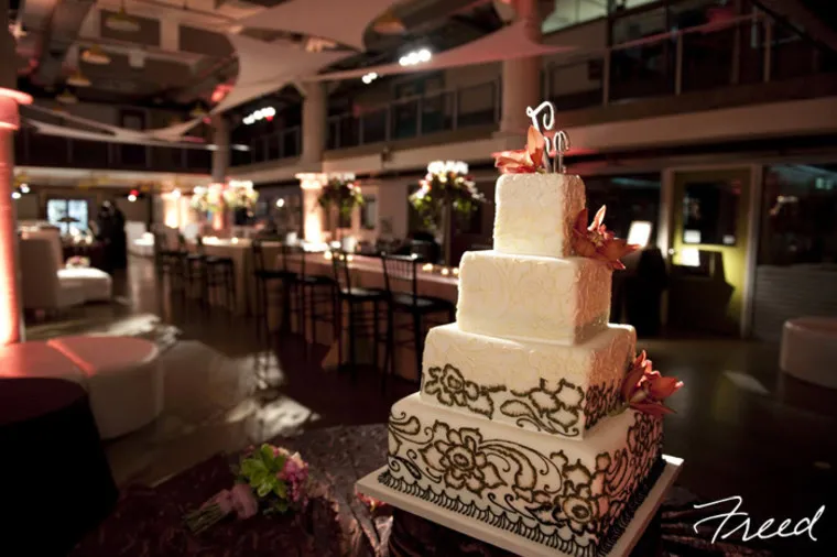 A three-tiered white wedding cake with decorative brown patterns and pink flowers sits on a table in an elegant, softly lit reception hall with tables, chairs, and floral centerpieces in the background.