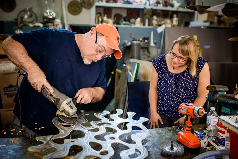 A man wearing an orange cap uses a power tool to shape a large abstract metal sculpture, while a woman in glasses and a blue polka-dot shirt watches attentively in a workshop filled with tools and materials.