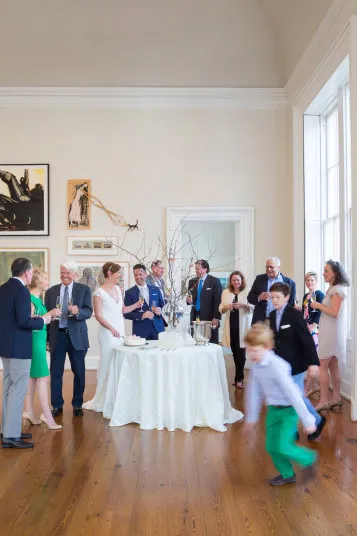 A group of people dressed formally gather around a round table with drinks in a bright room with large windows, while a child in green pants runs playfully in the foreground.