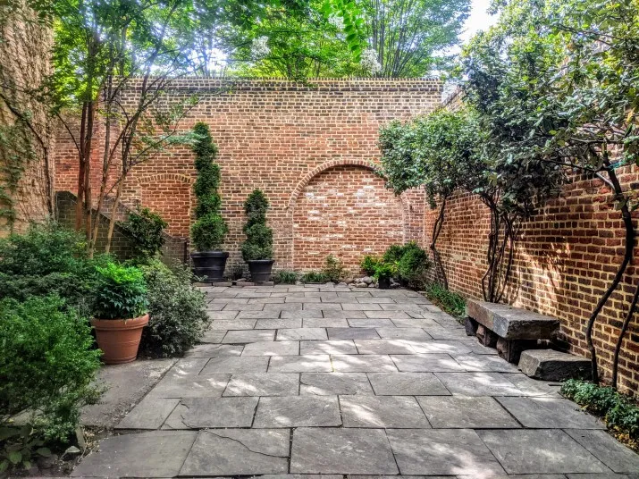 A peaceful courtyard with stone paving, surrounded by brick walls, potted shrubs, shaped topiary, lush green plants, and a stone bench under leafy trees. Sunlight filters through the foliage above.