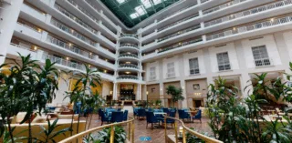 Spacious hotel atrium with multiple floors, white railings, a glass ceiling, and a central seating area with blue chairs, tables, and numerous green plants creating a modern, airy atmosphere.