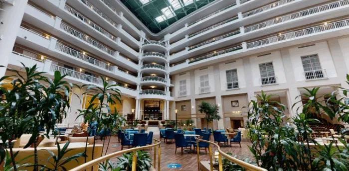 Spacious hotel atrium with multiple floors, white railings, a glass ceiling, and a central seating area with blue chairs, tables, and numerous green plants creating a modern, airy atmosphere.