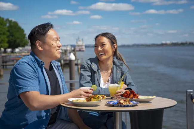 Two people sit at an outdoor table by the water, smiling and enjoying food and drinks. The weather is sunny with a blue sky and scattered clouds. Plates of food and beverages are on the table in front of them.