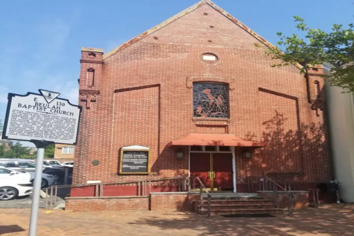 A red brick church with a large stained glass window above double doors, an orange awning, and a historical marker reading “Beulah Baptist Church” in front. Cars are parked to the left, and a tree shades part of the building.