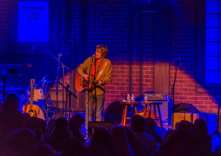 A musician plays an acoustic guitar and sings on stage under blue and purple lighting, with musical equipment and an audience visible. A sign reads Birchmere Stage Door on the brick wall backdrop.