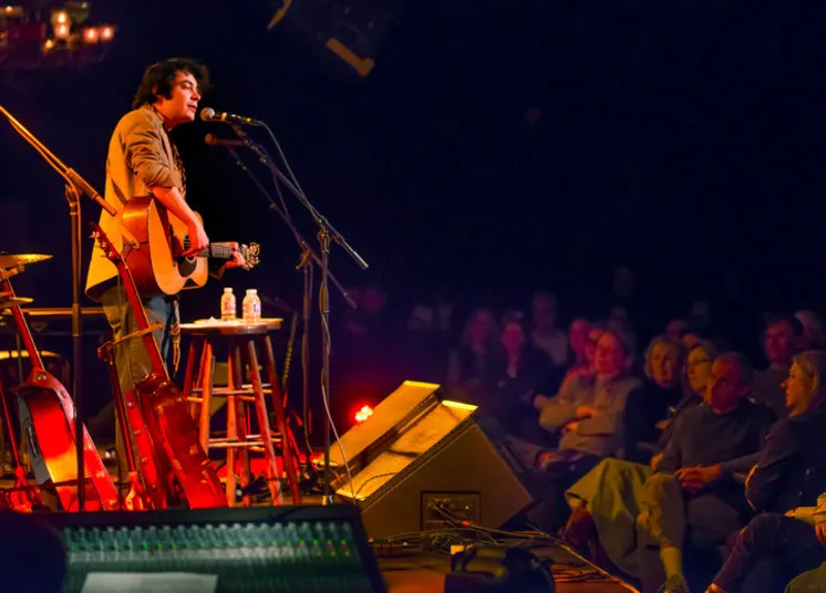 A musician plays an acoustic guitar and sings into a microphone on stage, lit by warm yellow and red lights, while seated audience members listen attentively in a dimly lit venue.