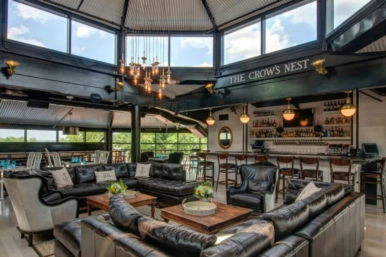 A stylish lounge with black leather sofas, wooden coffee tables, a chandelier, and a bar area with stools. Large windows and skylights provide natural light; The Crows Nest sign hangs above the bar.