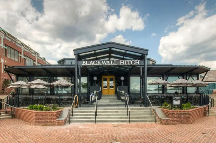 Front view of Blackwall Hitch restaurant with a glass entrance, outdoor seating on both sides with umbrellas, brick steps, and a partly cloudy sky above.
