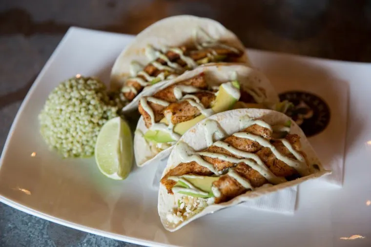 Three soft shell tacos filled with grilled fish, avocado slices, lettuce, and a creamy sauce, served with a wedge of lime and a side of cooked grains on a white plate.