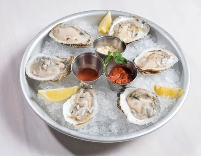 A round metal plate holding six raw oysters on half shells over crushed ice, with three lemon wedges and three small metal cups containing dipping sauces and garnishes.