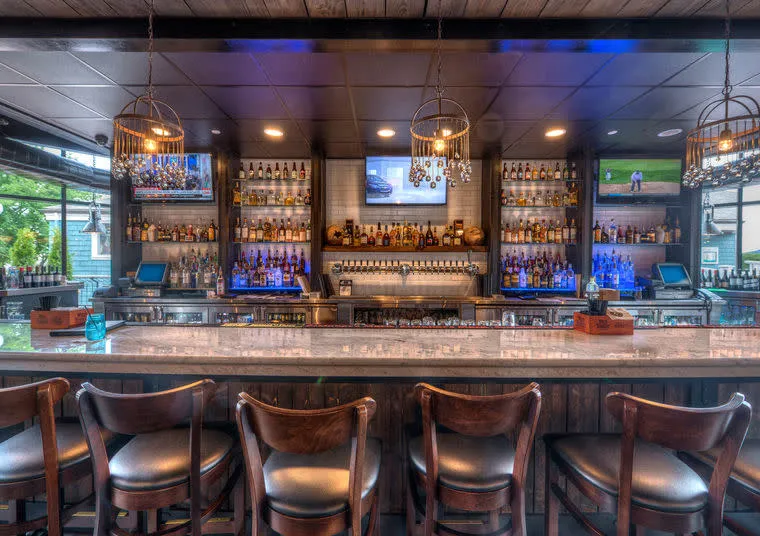 A modern bar with a marble countertop, five wooden chairs, shelves stocked with liquor bottles, hanging pendant lights, and flat-screen TVs displaying sports above the bar.