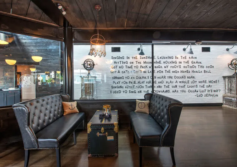 A cozy lounge with dark leather tufted sofas, nautical-themed pillows, and a trunk-style coffee table. Behind, a white brick wall displays large, handwritten Led Zeppelin lyrics in black text.