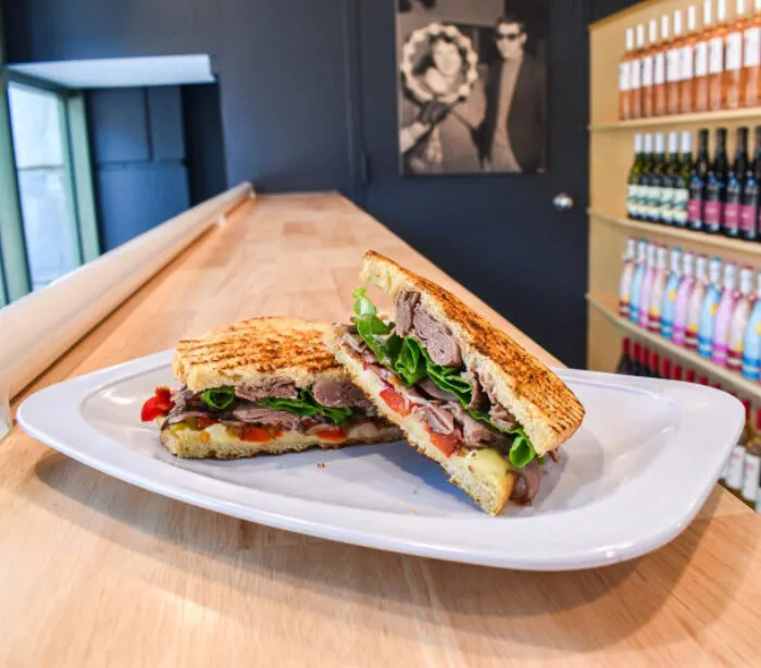 A toasted sandwich filled with roast beef, cheese, lettuce, and tomato is cut in half and served on a white plate atop a wooden counter, with shelves of bottled drinks and a black-and-white photo in the background.